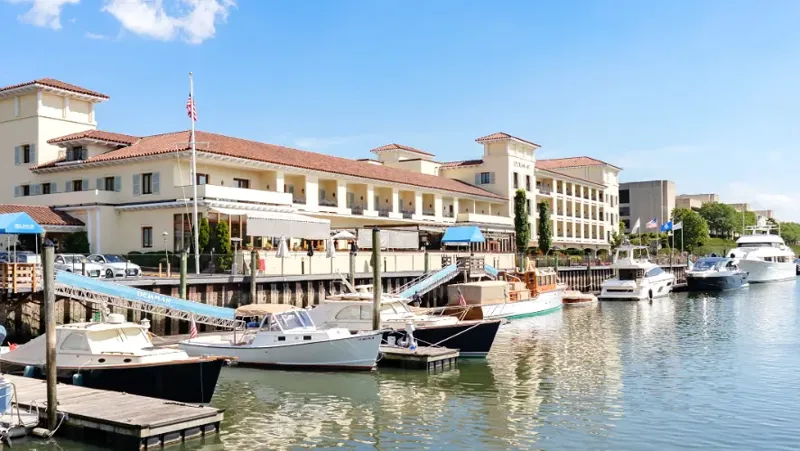 Waterfront marina scene featuring docked boats along a calm waterfront, adjacent to elegant multi-story buildings with terracotta roofs and large windows. Yachts and smaller vessels are moored, with clear skies enhancing the scenic coastal setting.