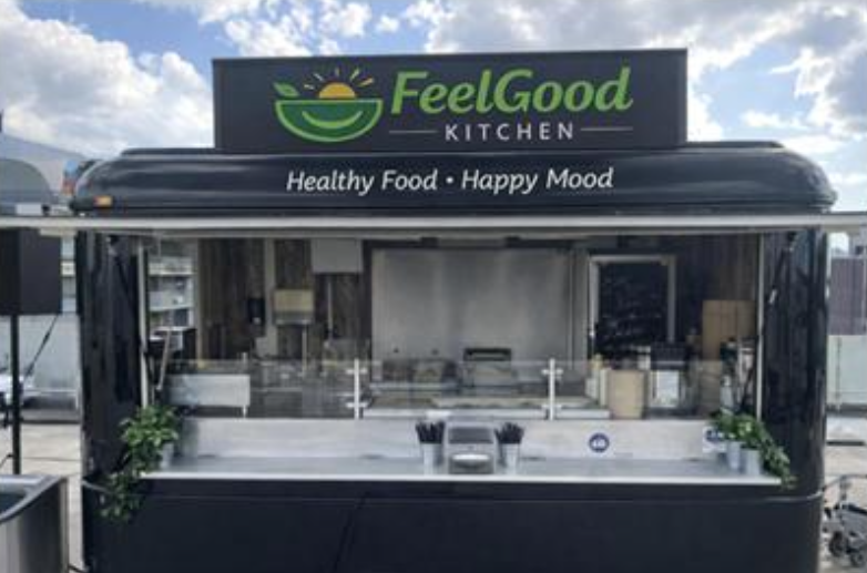 Mobile food truck branded FeelGood Kitchen displays healthy food and happy mood messaging. The setup includes a clean kitchen area with countertops, cooking stations, and a transparent window showcasing the event environment.