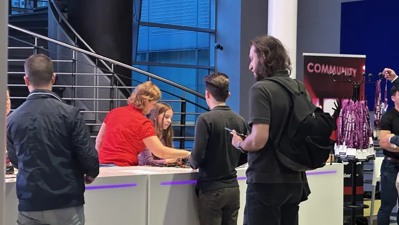 Attendees gather around a branded community booth at an indoor event, engaging in discussions at a table.