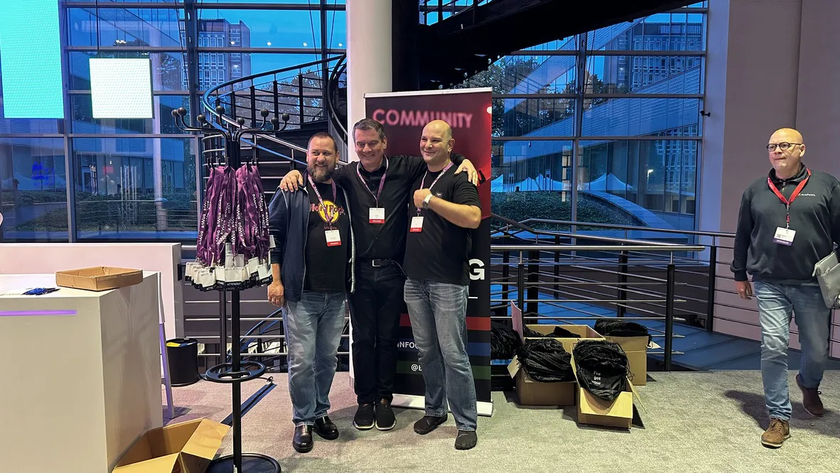 Three individuals pose at a branded community event booth featuring a sign with COMMUNITY text. The booth displays promotional items, including decorative scarves and branded merchandise, with open boxes of additional items in the background.