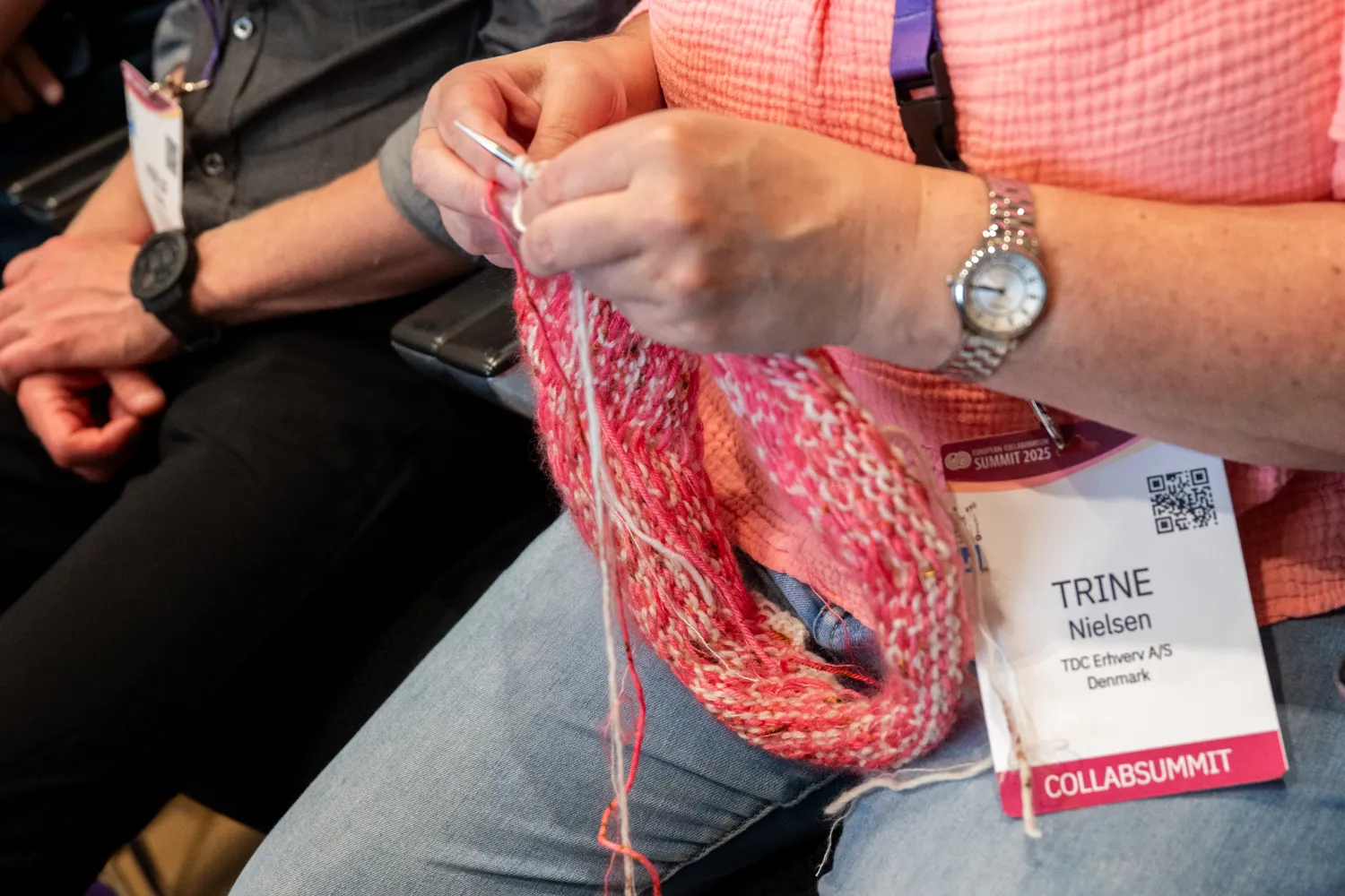 Attendee at a business conference knitting a colorful scarf, wearing a name badge indicating participation in an event. The scene captures an engaging, relaxed moment among professionals.
