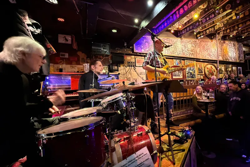 A lively band performs live music in a cozy, decorated venue with artistic wall displays. Attendees are seated and engaged, while musicians play electric guitar, drums, and sing into a microphone.
