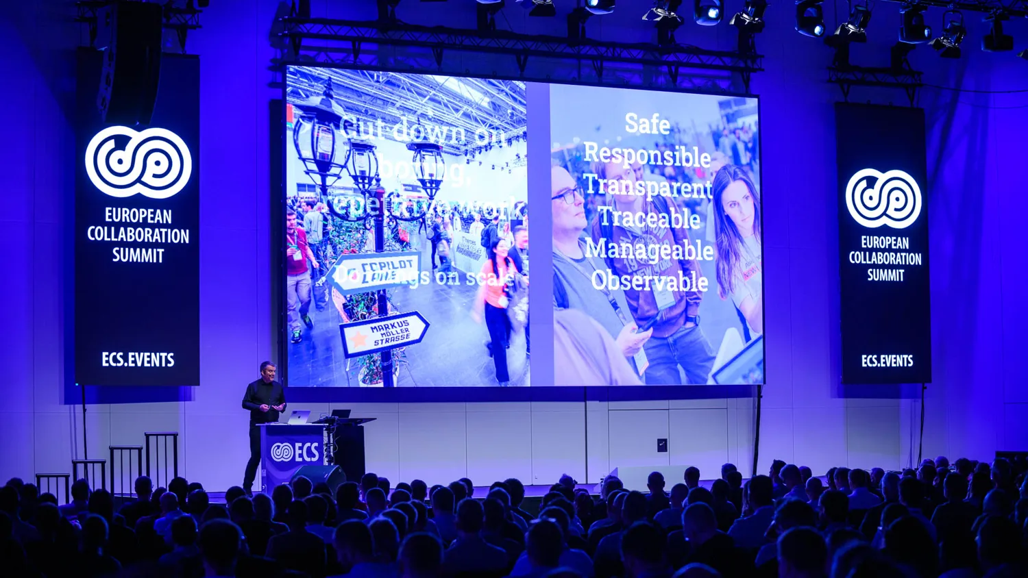 A speaker delivers a presentation at the European Collaboration Summit, flanked by large screens displaying event branding and key themes such as safety, traceability, and responsible practices. The audience attentively faces the stage with illuminated blue lighting.