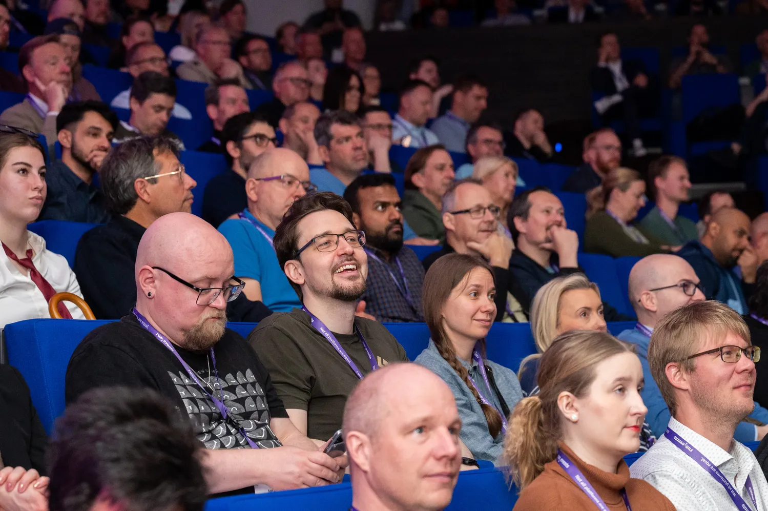 Professional audience attentively seated in blue rows during a conference session, engaging with the presentation. Attendees wear lanyards and exhibit varied expressions of interest and focus.