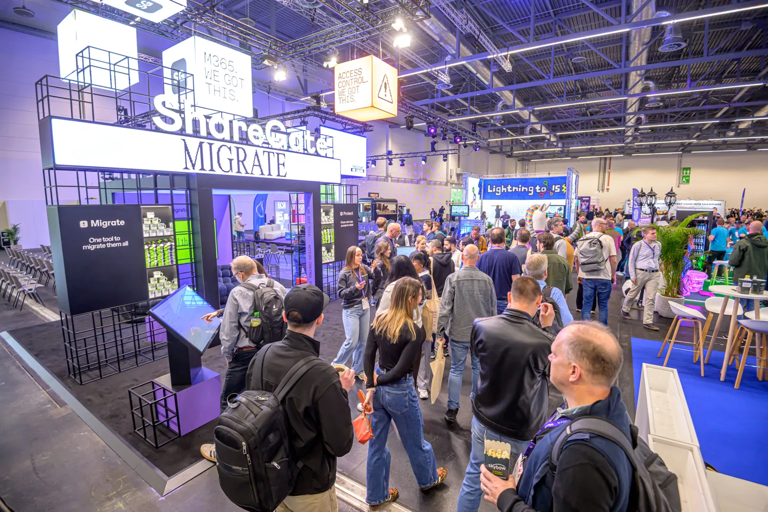 Professional attendees gather around a tech-focused booth promoting digital migration solutions. The booth features illuminated signage, interactive displays, and branded promotional materials in a bustling exhibition hall.