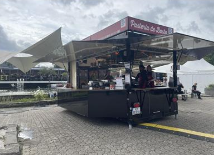Mobile pastry vendor stall labeled "Pastería de Basilio" serving food at an outdoor event venue, featuring modern architectural structures in the background. Staff are visible behind the counter preparing and serving pastries and baked goods.