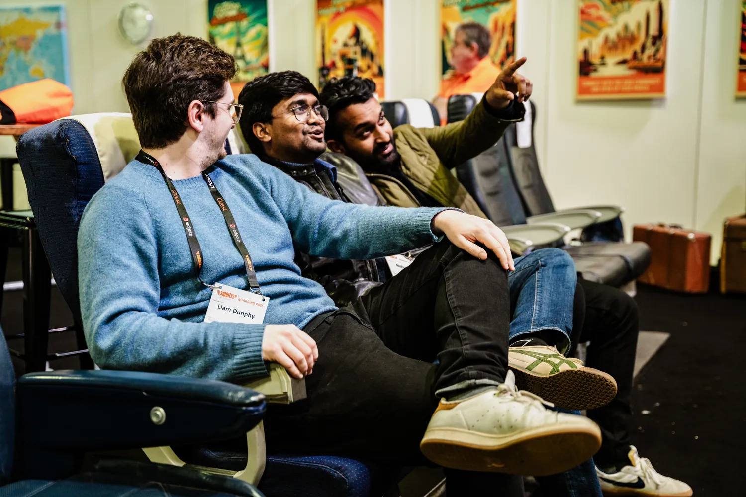 Three individuals seated in office chairs at what appears to be a conference or networking event. They are engaged in conversation, with one gesturing while wearing casual business attire and lanyards. Vibrant artwork adorns the wall behind them.
