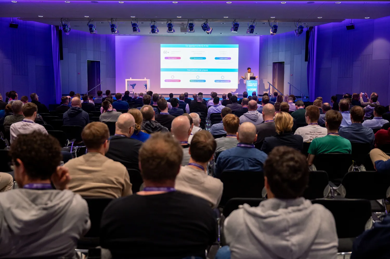 A professional audience attentively listens to a speaker delivering a presentation at a conference venue. The stage features an illuminated screen displaying data visualizations and statistics, while attendees are seated facing forward.
