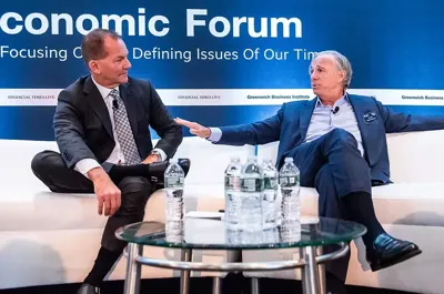 Two men engaged in a professional discussion at an economic forum. They are seated on a stage, gesturing with hands, and positioned in front of branded backdrop displaying "Economic Forum" and "Greenwich Business Institute." Water bottles are placed on a transparent tabletop between them.