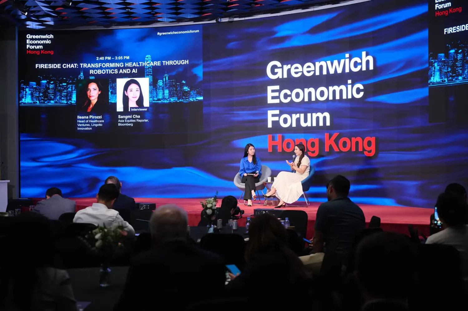 Two professionals engaged in a panel discussion titled "Transforming Healthcare Through Robotics and AI" at the Greenwich Economic Forum in Hong Kong. The event features a stage backdrop with event branding, city skyline imagery, and audience members attentively observing the conversation.
