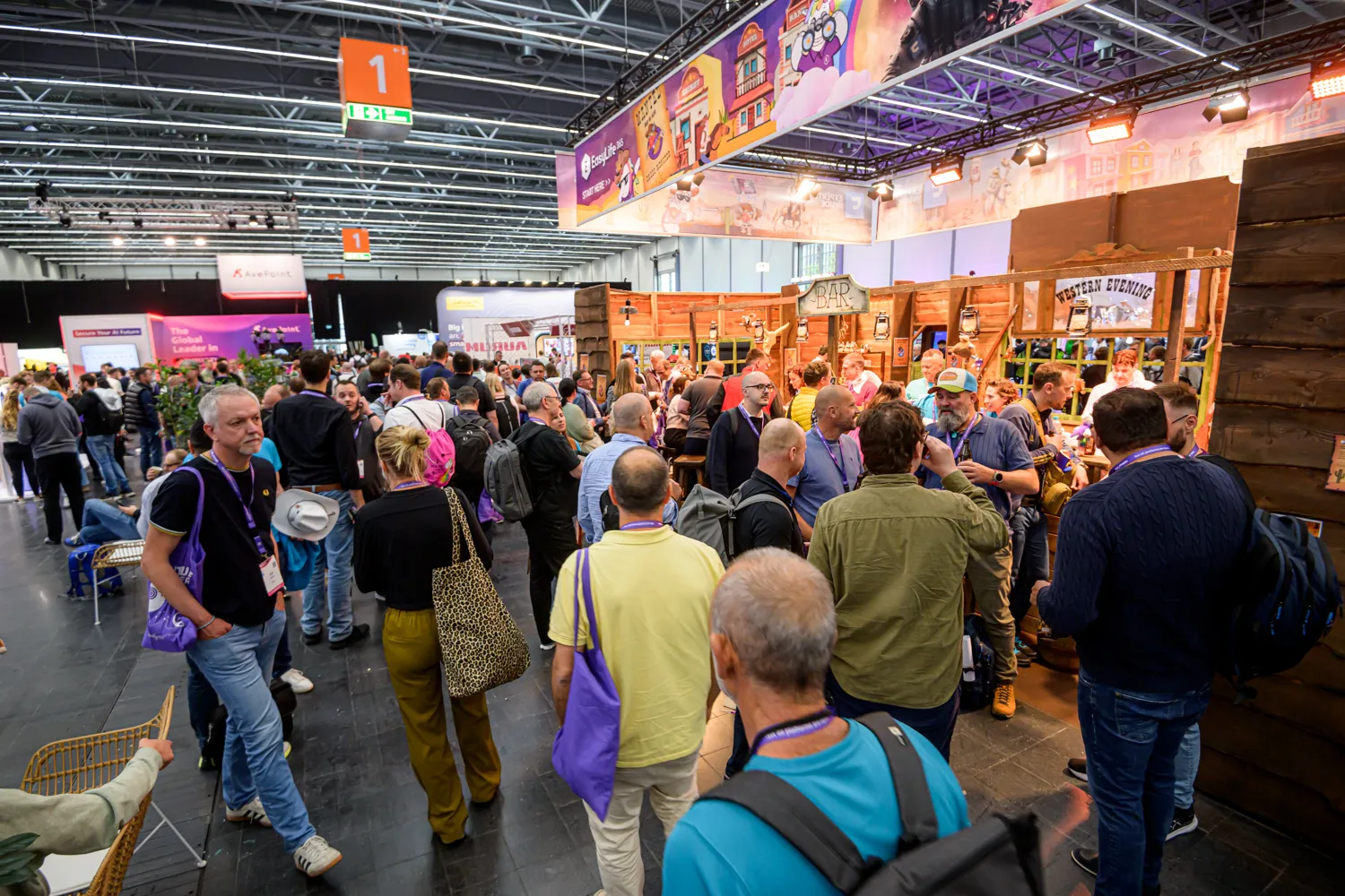 Attendees gather around a rustic-themed exhibition booth at a busy indoor trade event, engaging with displays and each other. The scene features vibrant signage, interactive elements, and diverse participants in casual business attire.