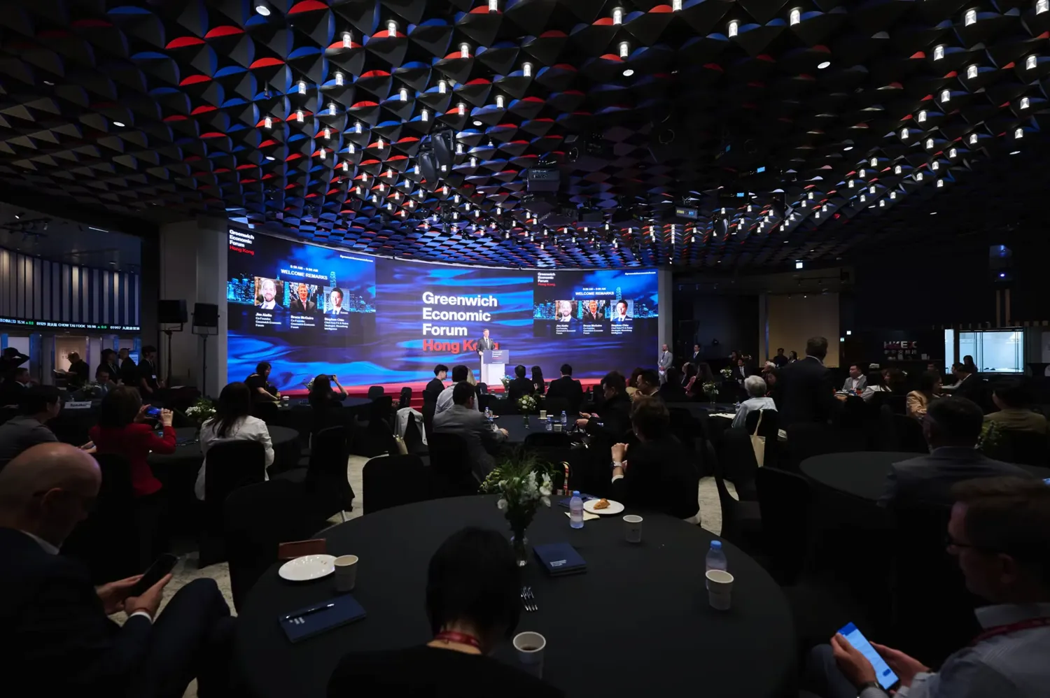 Professional conference attendees seated at tables in a modern venue, facing a stage with speakers and a large screen displaying the "Greenwich Economic Forum Hong Kong" branding. The event features geometric ceiling lighting and participants engaged in discussions, capturing an organized business forum atmosphere.