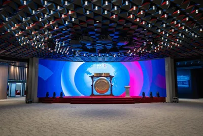 A grand conference stage features a vibrant, illuminated backdrop with geometric patterns and a central podium. The scene includes seated attendees and a prominent speaker area with digital display elements.