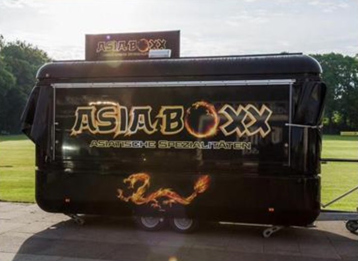 Black food truck branded with "AsiaBoxx" featuring bold, fiery typography and a stylized dragon design. The truck advertises Asian specialties and is positioned outdoors on a grassy area during daylight.