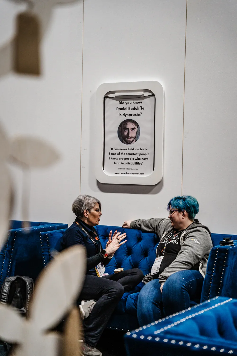 Two individuals engaged in conversation on a blue seating arrangement during an event. Behind them, a framed display highlights actor Daniel Radcliffe discussing neurodiversity and learning disabilities.