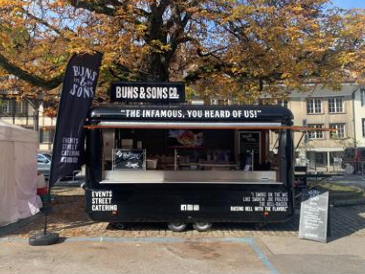 Mobile food truck branded Buns & Sons Catering displays its offerings at an outdoor event, featuring signage advertising menu items and catering services. The setup includes a branded banner, promotional text, and a chalkboard menu in an urban outdoor setting.