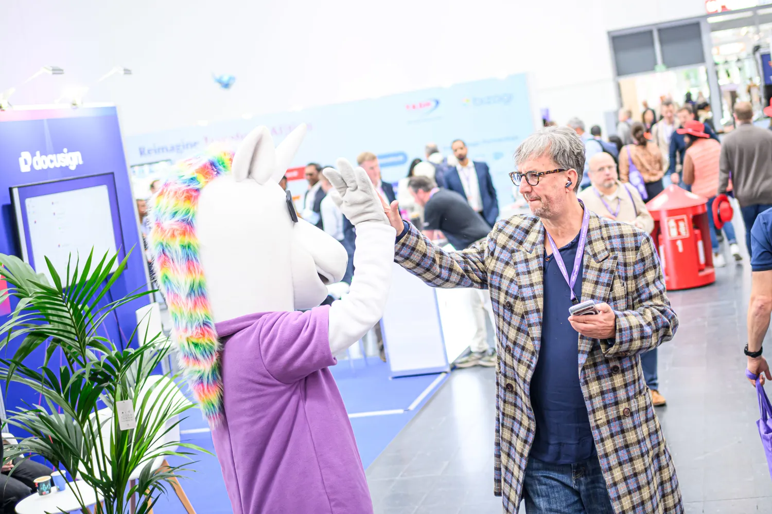 Attendees at a professional trade show booth featuring a person interacting with a colorful, whimsical mascot unicorn prop. The scene includes participants in business attire, an informational display board, and decorative plants.