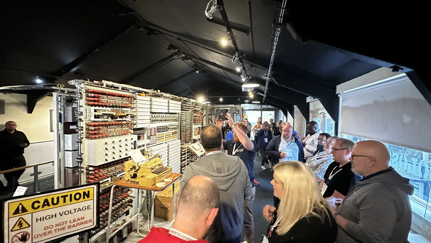Group of people observing an intricate high-voltage technical exhibit with complex wiring and components. Attendees are engaged, with safety warnings prominently displayed indicating caution due to high voltage.