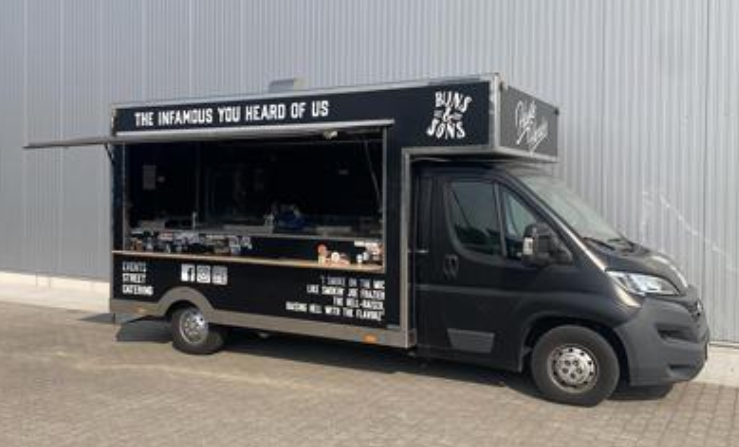A black food catering truck displays branding for "The Infamous You Heard Of Us" and "Cafe Jones." The vehicle features an open food preparation area with various menu items visible, positioned outdoors near a building.