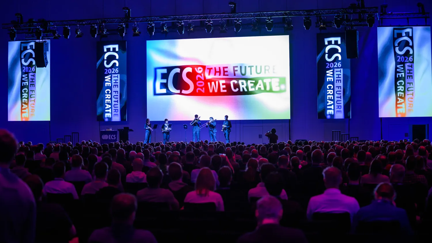 A large audience attends an event featuring a stage presentation with a vibrant backdrop displaying "ECS 2026: The Future We Create." Speakers and musicians perform on the stage, illuminated by dramatic lighting.