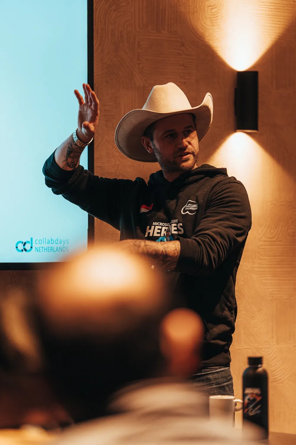 Man in cowboy hat presenting or speaking at a conference setting, wearing a branded hoodie with "Microsoft Heroes" and "Azure" logos. The backdrop displays "CollabDays Netherlands" branding.