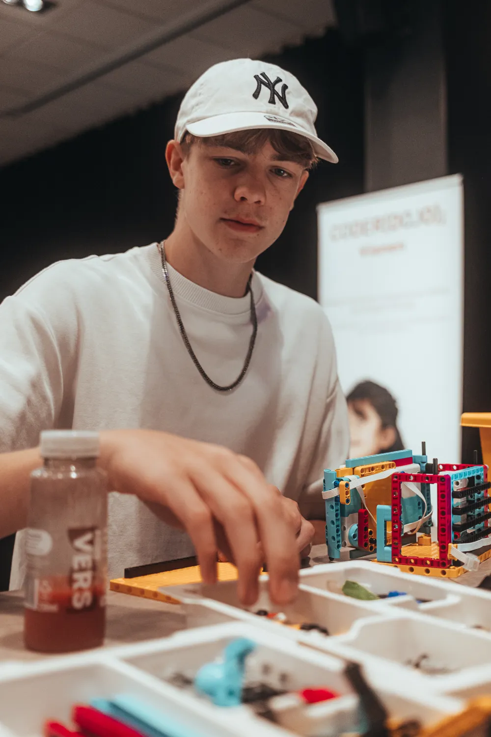 A person wearing a cap and light-colored shirt is assembling a colorful robotic mechanism using interlocking blocks. The individual holds a beverage can and appears to be engaged in a hands-on engineering or STEM activity.