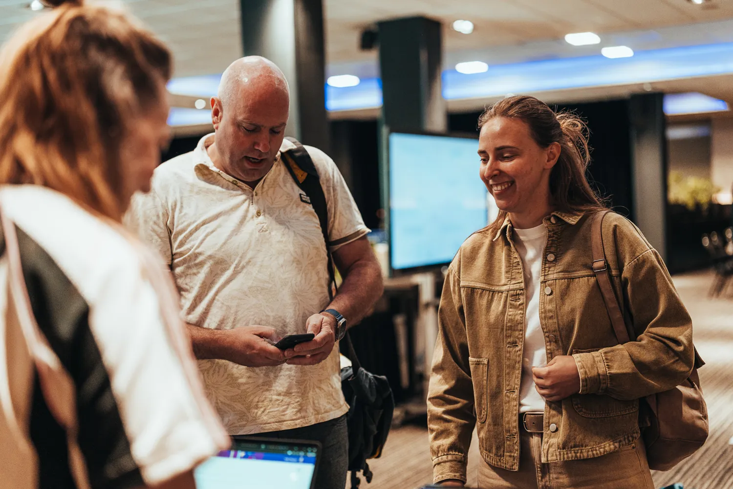 Three individuals in a modern indoor setting appear to be engaged in a discussion. A man checks his phone while two others, one in casual attire and another in a denim jacket, smile and interact warmly. The background includes lighting fixtures and a display screen.