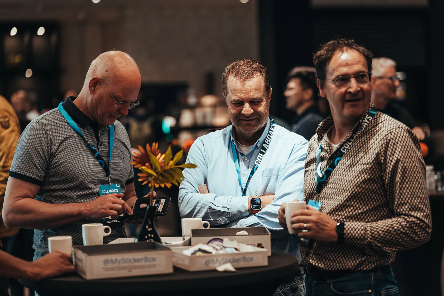 Three attendees at a conference booth exchange items and engage in conversation. The scene features branded lanyards, promotional merchandise boxes, and a relaxed, collaborative atmosphere in a well-attended event setting.