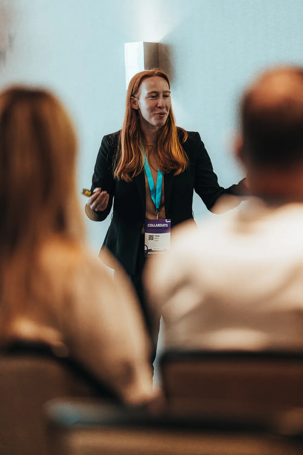 A professional presenter in a business attire stands at a podium, addressing an audience at a conference. The individual holds a microphone and wears a lanyard with event identification badges, indicating participation in a formal networking or business event.