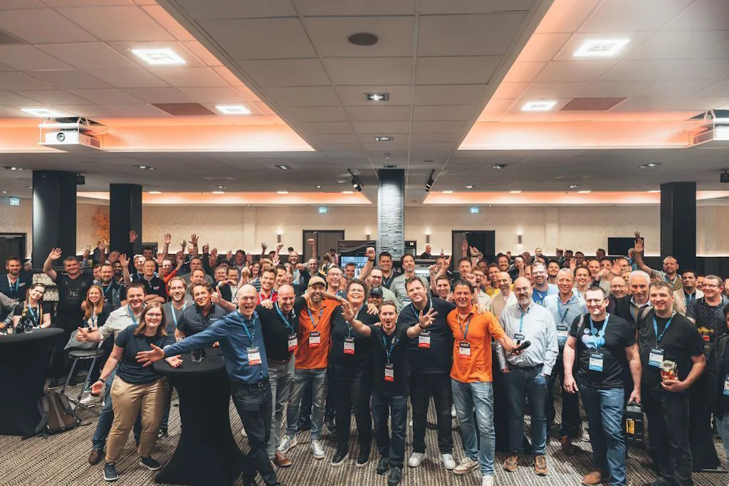 Attendees at a professional conference pose together in a spacious conference hall, wearing event badges and casual business attire, exuding enthusiasm and camaraderie. The setting features modern lighting and seating arrangements in the background.