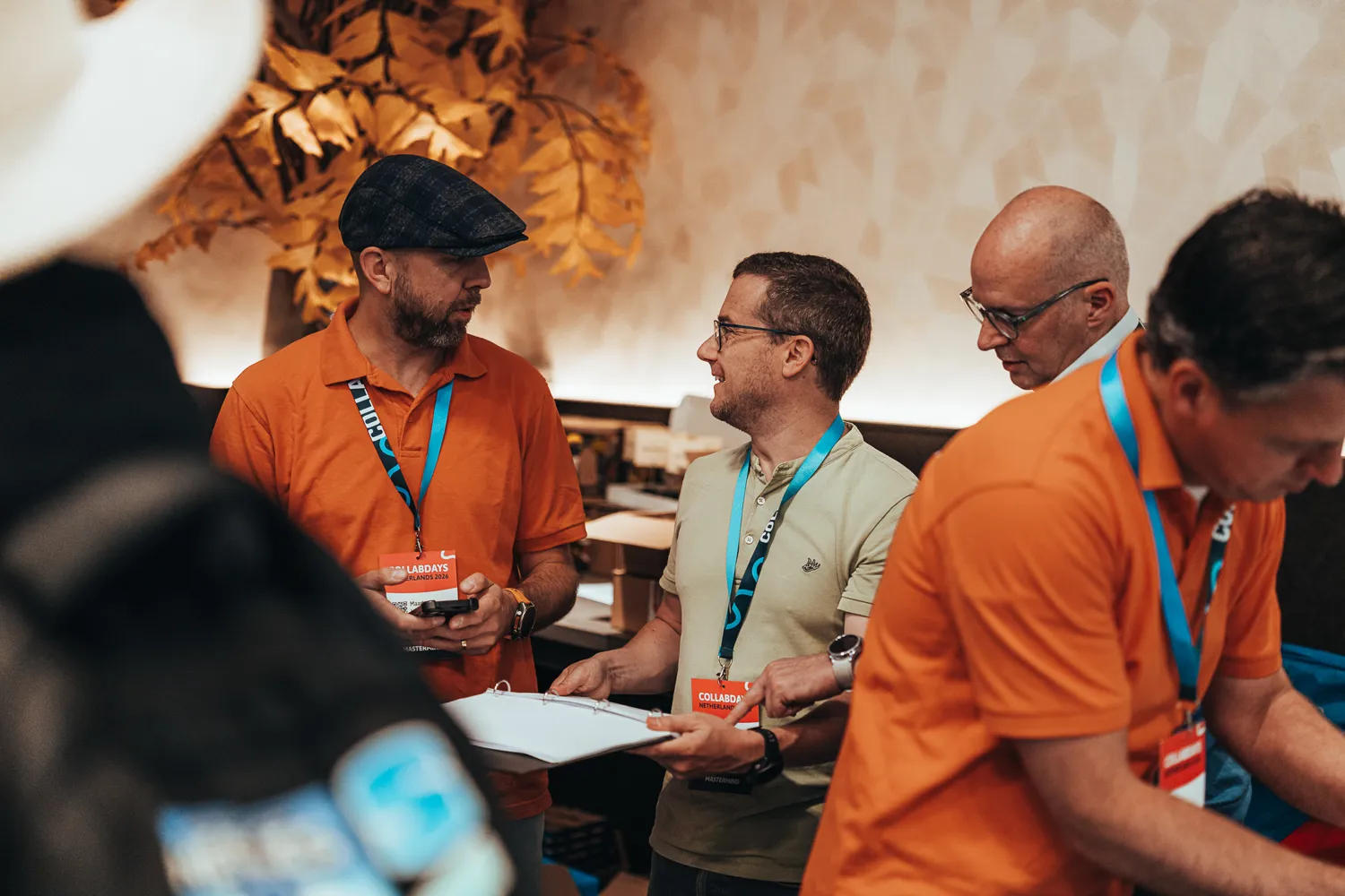 Attendees in an indoor event space engage in discussion, wearing branded lanyards and orange shirts. The background features decorative, textured golden foliage elements.