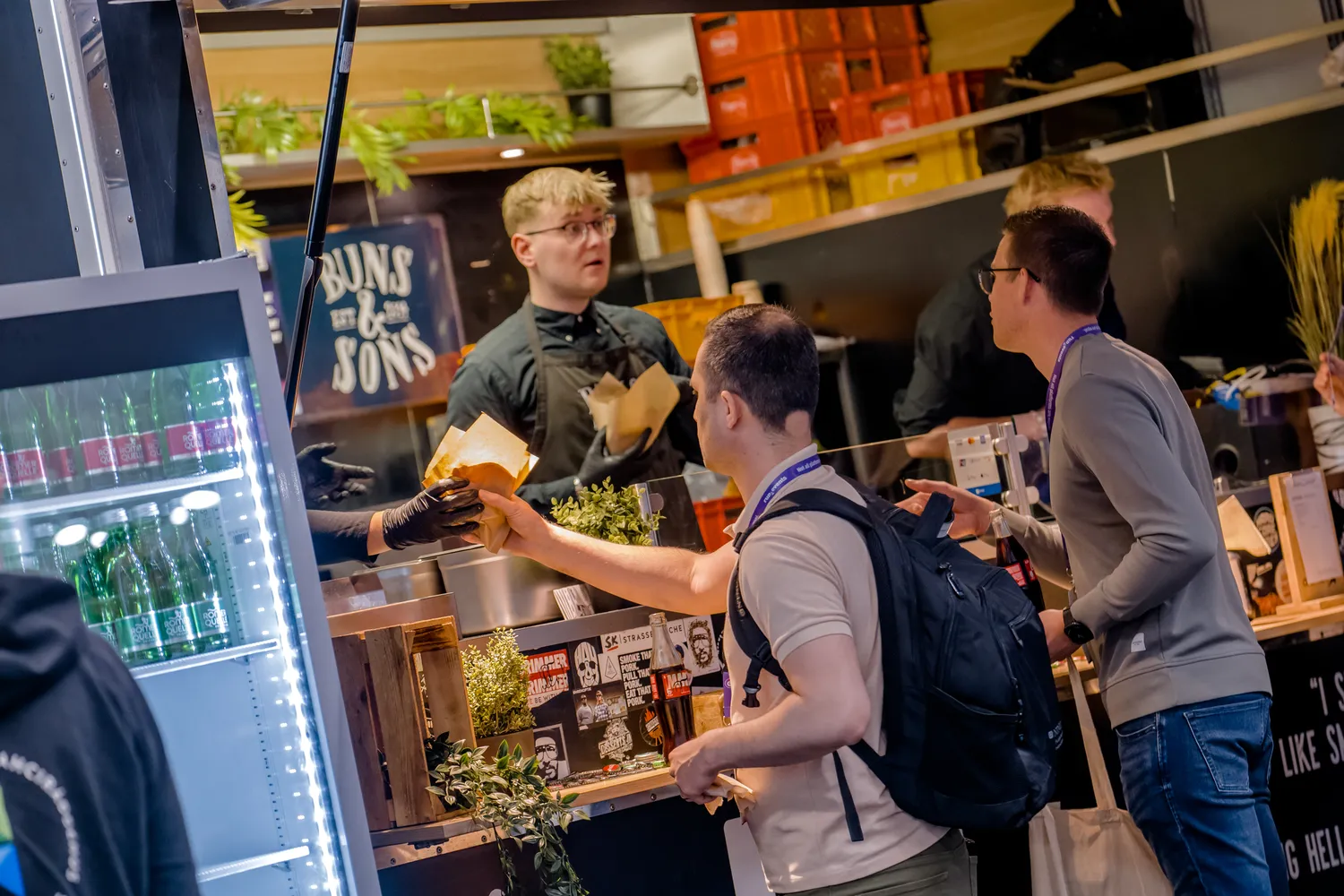 Attendees at a lively event interact with a food vendor booth, receiving fresh greens in takeaway containers. The setting features vibrant decor with shelves displaying branded merchandise and plants, creating an engaging and casual atmosphere.