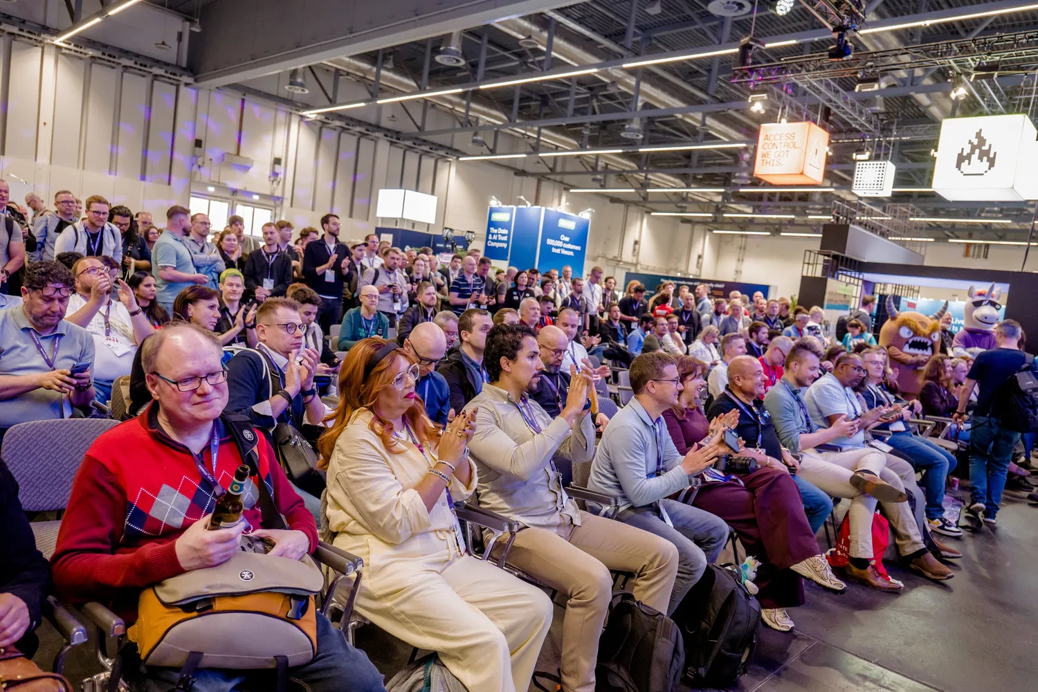 A lively professional conference audience seated in rows, clapping enthusiastically during a presentation. Attendees wear business attire, lanyards, and some access badges visible in the background.