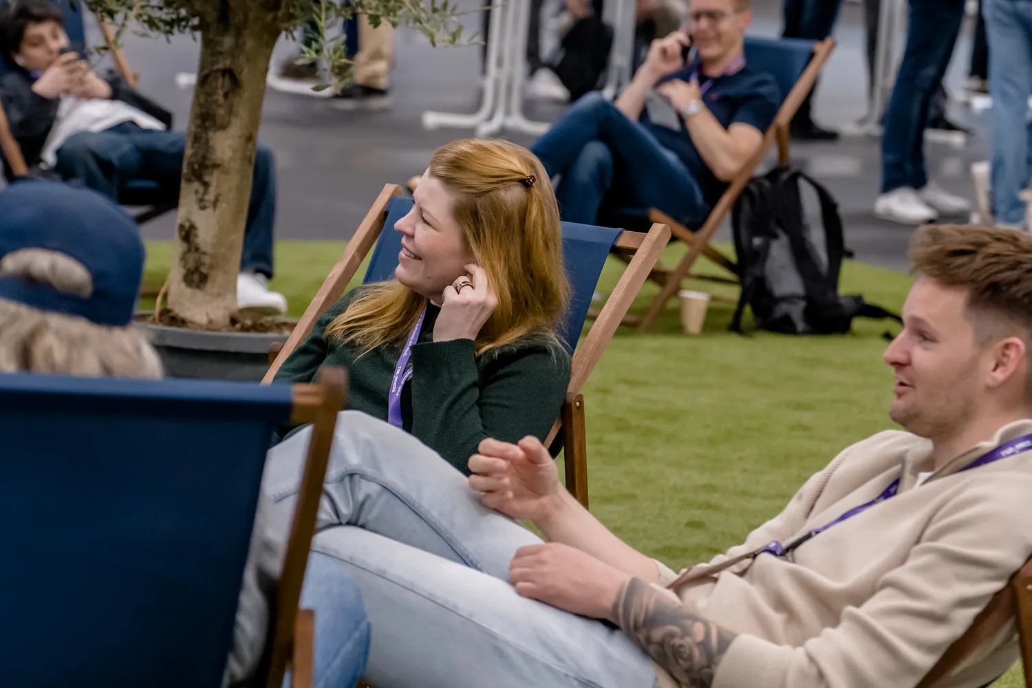 Attendees at an outdoor event are seated on lounge chairs, engaging in conversation. The scene includes individuals wearing lanyards, some taking part in discussions while others appear to be on a phone call.