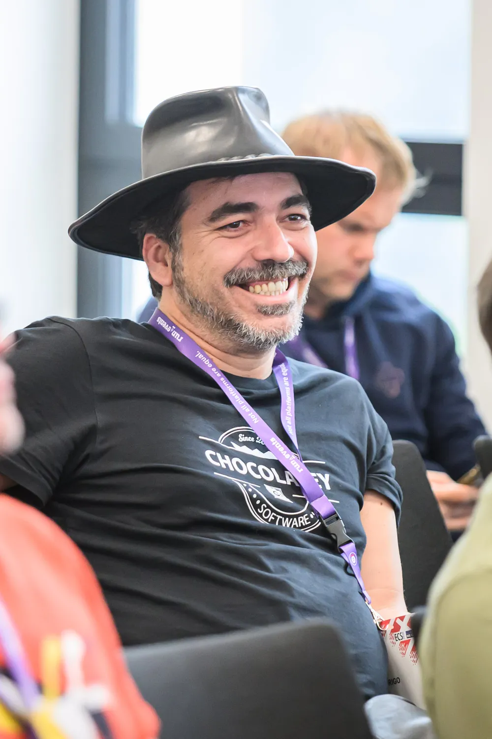 A man wearing a black cowboy hat and dark shirt with "Chocolatey Software" branding smiles warmly. He is attending an event, identifiable by a purple lanyard with event branding.