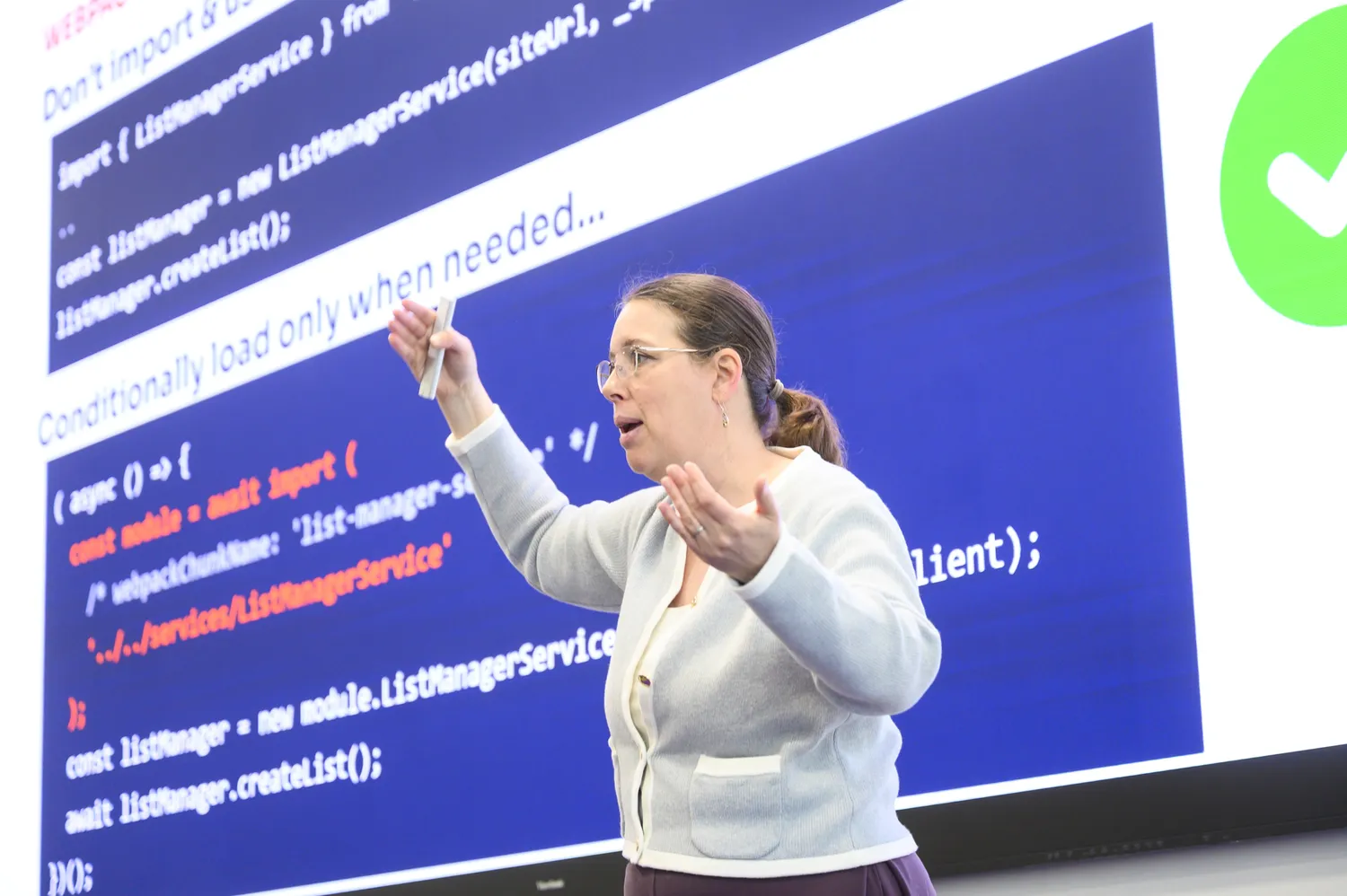 A woman presents technical concepts using hand gestures while explaining code snippets on a digital screen filled with programming syntax. She appears engaged in an educational or conference setting discussing software development.