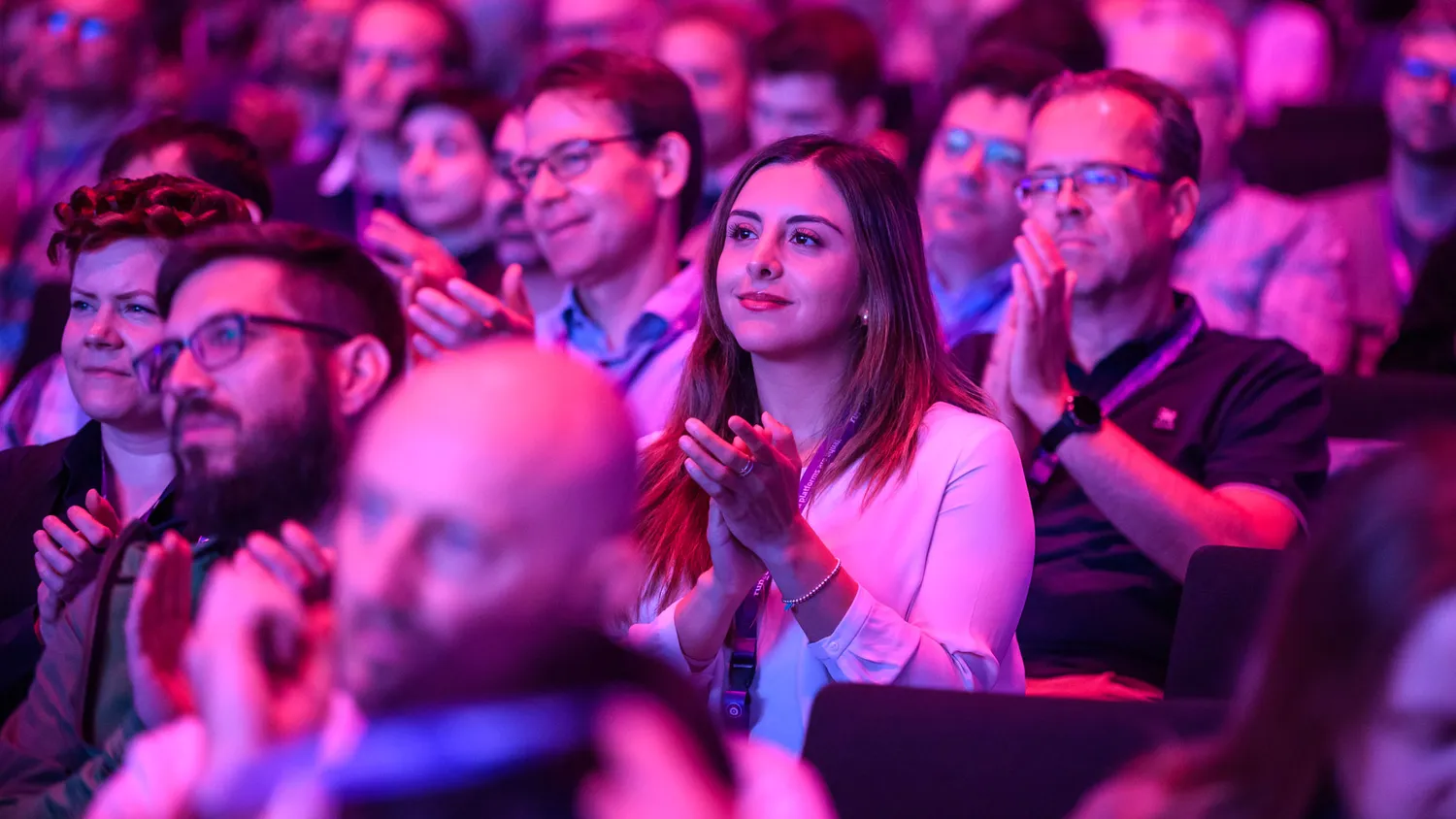 Attendees at a professional event clap enthusiastically during a presentation, with a woman in the foreground clapping her hands. The scene features an engaged audience under vibrant stage lighting.