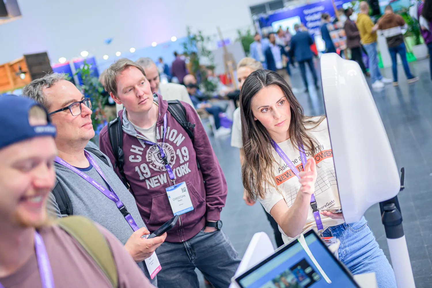 Attendees at a professional conference gather around a large interactive whiteboard display. A presenter points to content, while participants in business attire and event badges appear engaged in discussion.