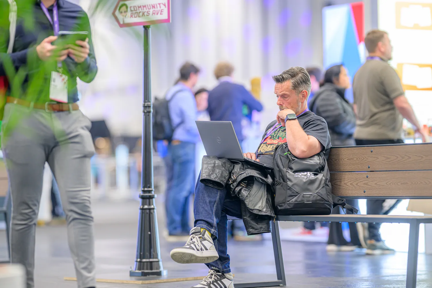 A man sits on a bench using a laptop during what appears to be an indoor event. He is surrounded by other attendees, with colorful signage and directional markers in the background.