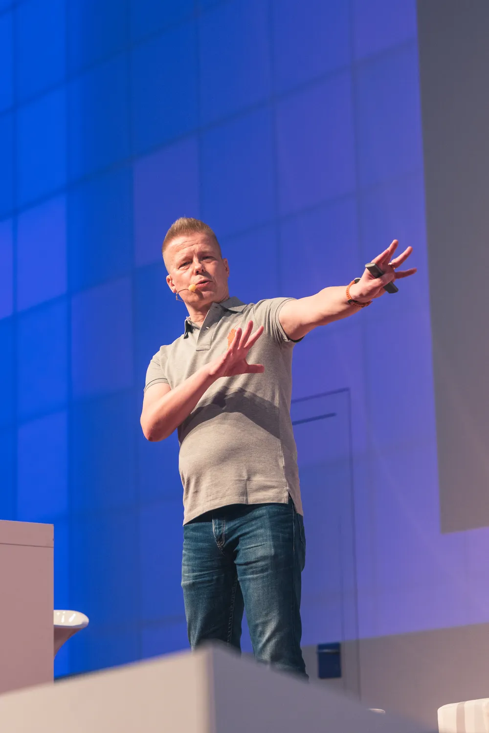 A man stands on a stage delivering a presentation, gesturing with both hands to emphasize points. He is dressed in casual business attire against a modern, illuminated backdrop.