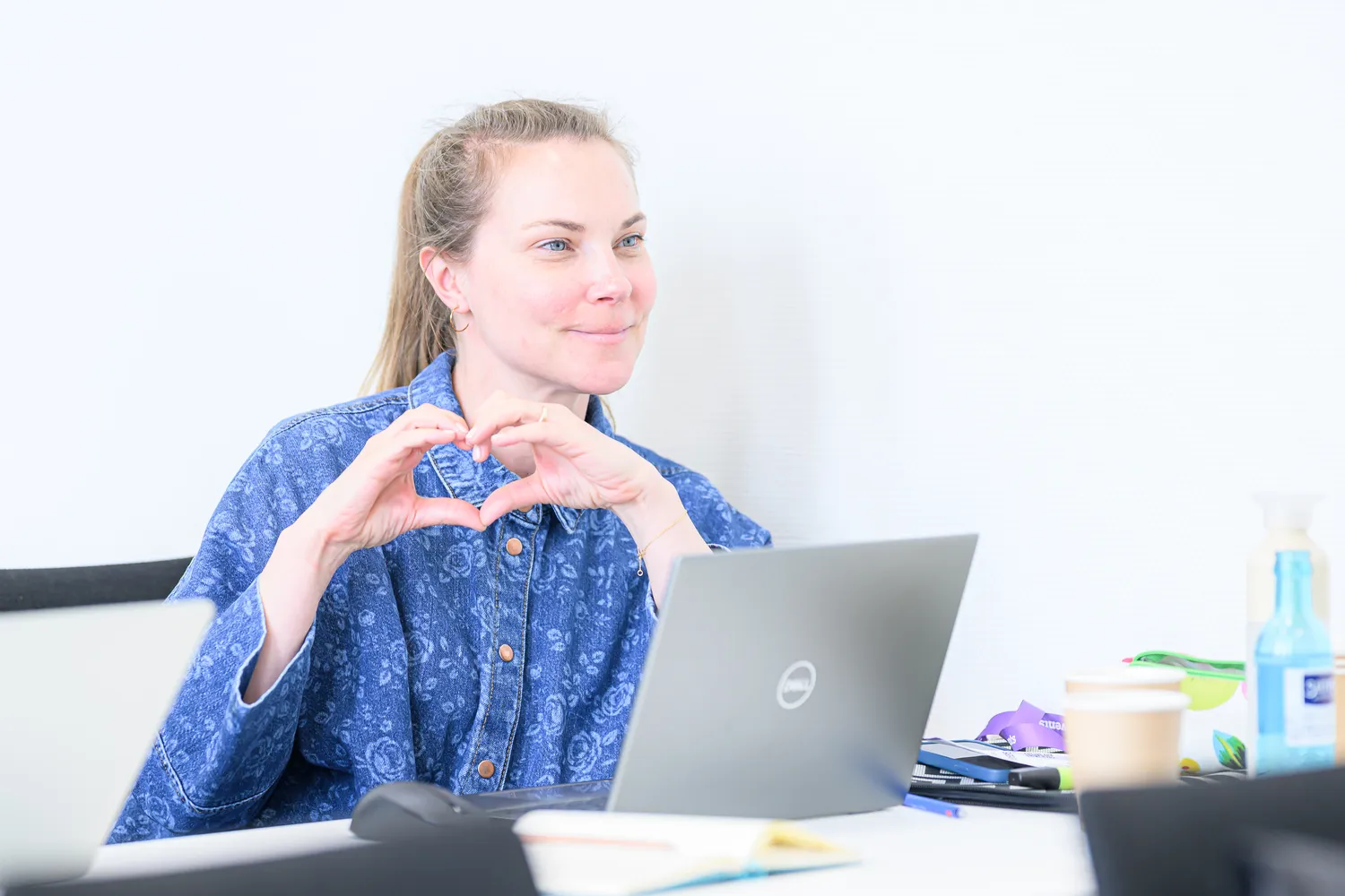 A person wearing a patterned shirt forms heart shapes with their hands while seated at a home office desk. The scene includes an open laptop, office supplies, and beverages on the desk.