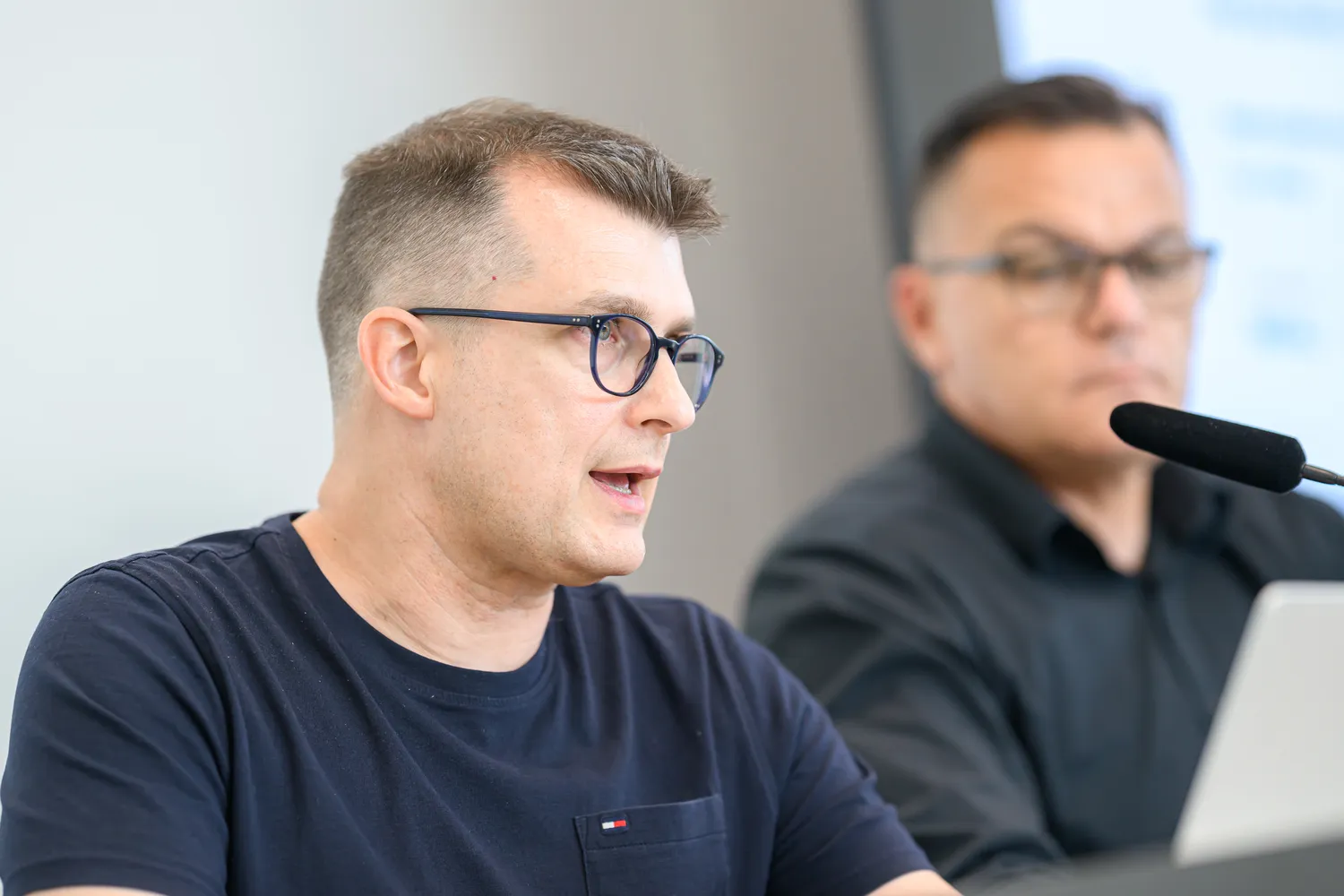 A man in glasses and a casual shirt speaks into a microphone at what appears to be a professional conference or panel discussion. Another individual, partially visible, is also speaking from the podium.