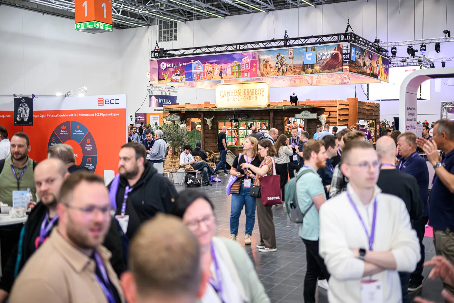 Professional trade expo attendees gather at a booth with western-themed decor, engaging in discussions and networking. Visible branding includes booths from companies like BCC and prominent overhead signage featuring visuals and text.