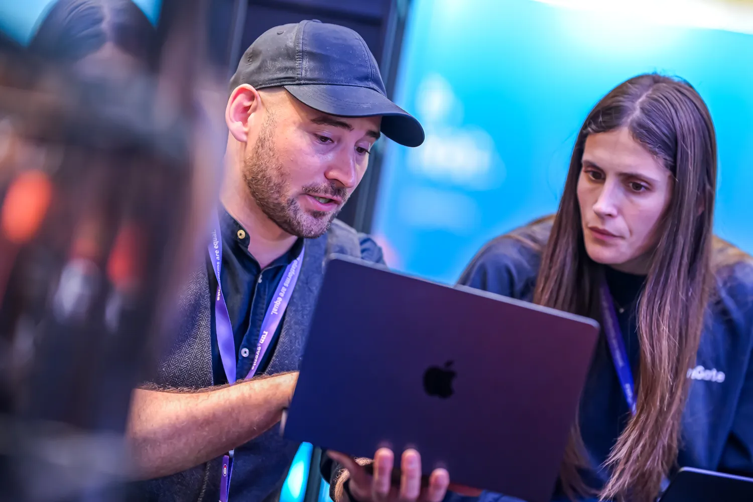 Two individuals engaged in a discussion at a tech conference, examining an Apple MacBook. The setting appears vibrant with blurred attendees in the background.
