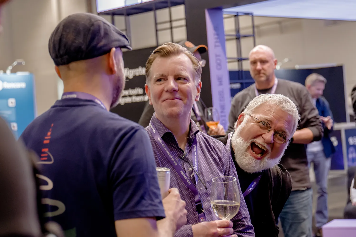 Group of attendees at a professional conference booth engaging in conversation and toasting with wine glasses. The scene captures lively interaction among participants in a well-lit indoor setting with branded event attire and promotional displays.