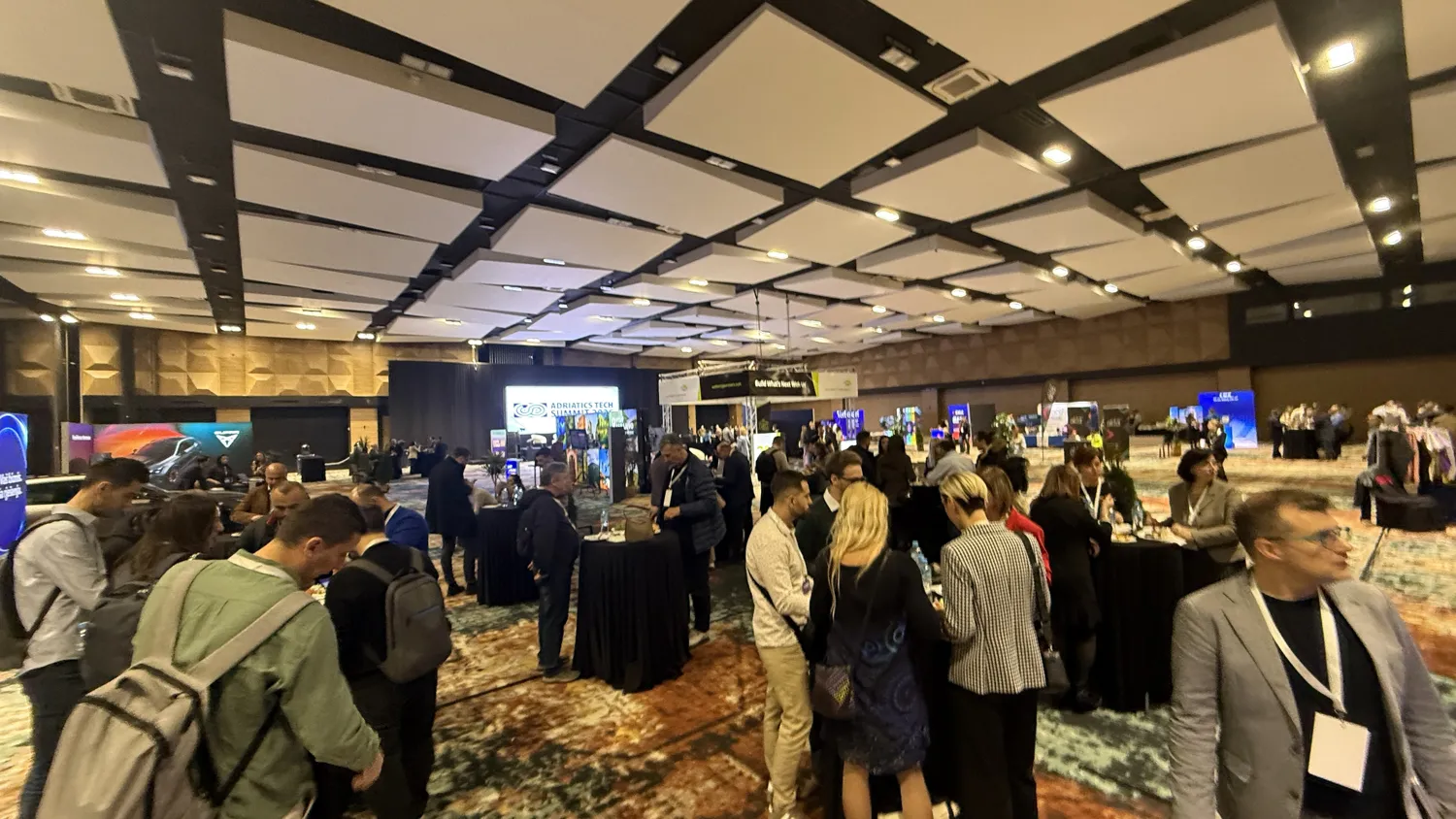 Professional attendees gather at a bustling trade show booths area with modern exhibition hall lighting. Visible are branded displays, interactive presentations, and participants engaging in discussions and networking.