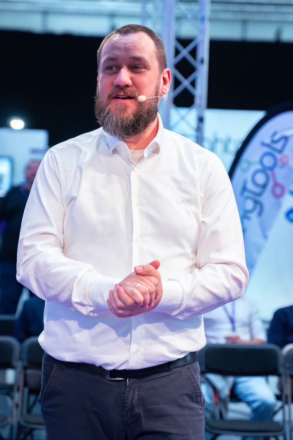 Man in a white patterned shirt and dark pants speaking at an indoor event, gesturing with his hands while wearing a microphone clip. The background shows seating arrangements and stage lighting.