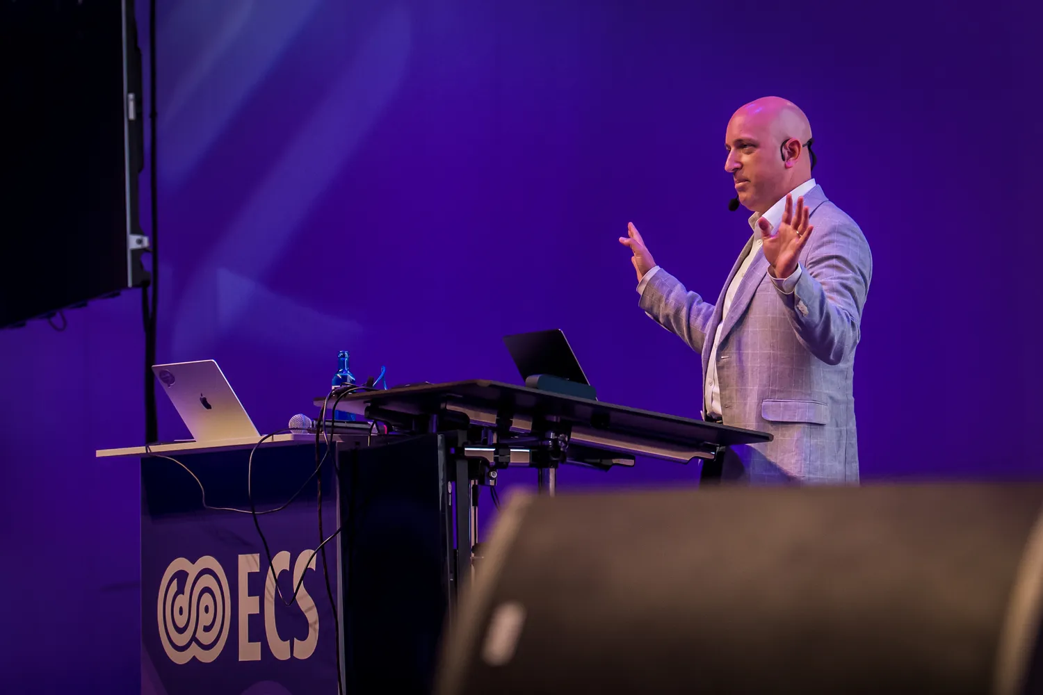 A man in a suit stands on a stage delivering a presentation with hand gestures, using a podium that holds electronic devices. The backdrop features the logo of an organization, and an audience section is partially visible.