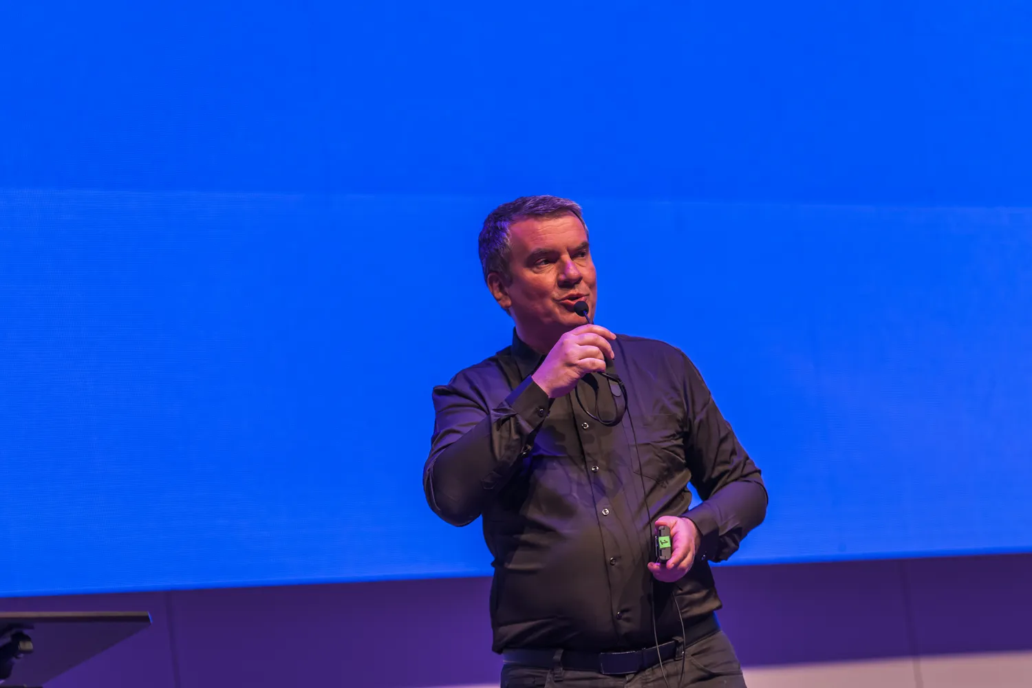 Man in professional attire stands on a stage, speaking into a microphone while holding a small device, likely for presentation slides. The background features vibrant stage lighting with blue and purple tones.
