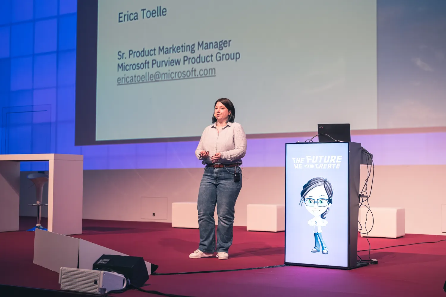 A woman presenting at a conference stage displays her name, title as Senior Product Marketing Manager from Microsoft Purview Product Group, and contact email. She stands behind a branded podium with an animated character graphic and text "The Future We Create" alongside audio-visual equipment.